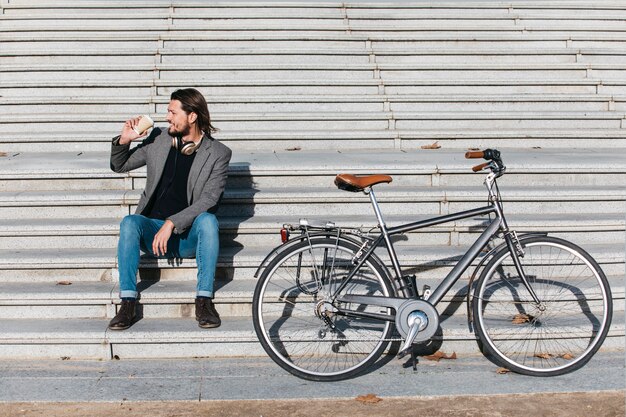 Elegante jovem sentado na escadaria com bicicleta bebendo café