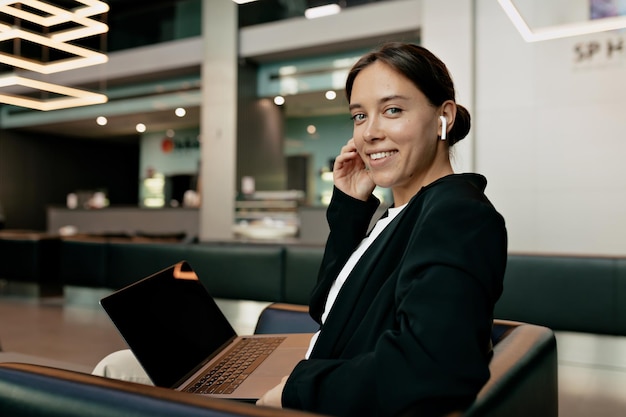 Elegante jovem trabalhadora de terno escuro em fones de ouvido está segurando laptop e sorrindo para a câmera enquanto trabalha Mulher muito atraente está trabalhando com laptop no escritório moderno