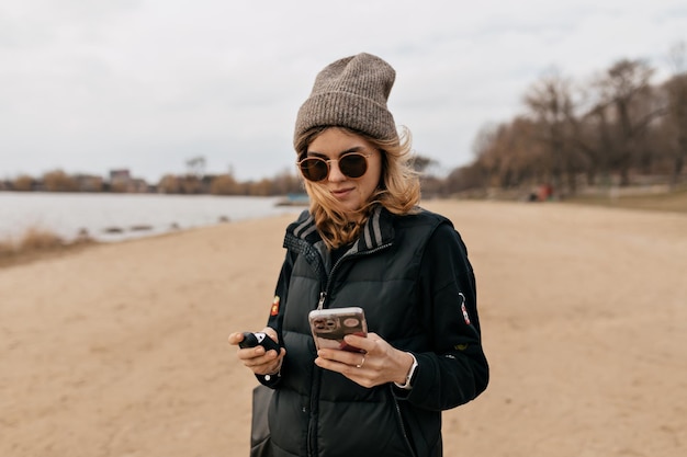 Elegante jovem usando óculos escuros e boné está rolando smartphone na praia em dia quente e ensolarado de primavera
