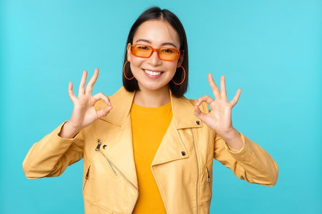 Elegante linda mulher asiática em óculos de sol sorrindo espantado mostrando sinal ok ok recomendando smth suporta excelente escolha em pé sobre fundo azul
