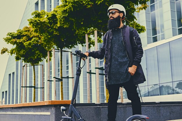 Elegante macho barbudo em óculos de sol posando na scooter elétrica sobre fundo de construção moderna.