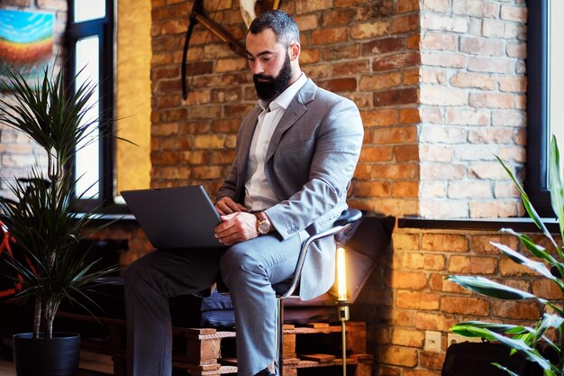 Elegante macho barbudo trabalha com um laptop em uma sala com interior loft.