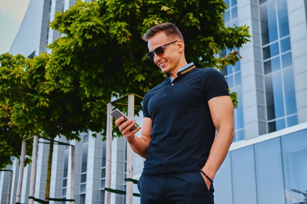 Elegante macho musculoso usando smartphone sobre fundo de construção moderna no centro da cidade.