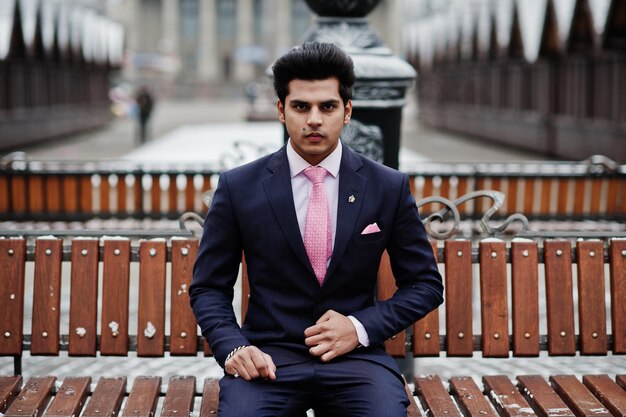 Elegante modelo de homem macho indiano de terno e gravata rosa posou no dia de inverno e sentado no banco