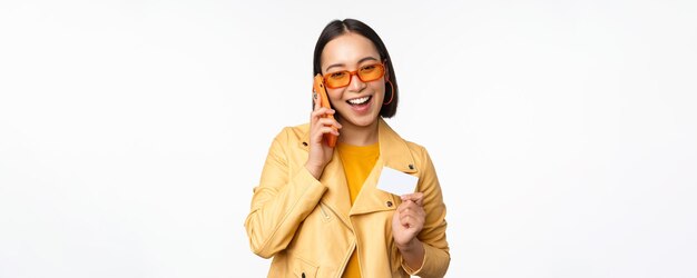 Elegante modelo feminino asiático falando no smartphone e mostrando o cartão de crédito sobre fundo branco