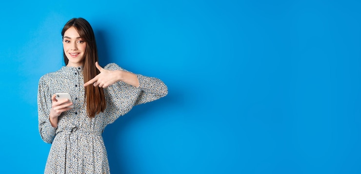 Foto grátis elegante mulher caucasiana vestida apontando para smartphone e sorrindo mostrando promoção online em pé