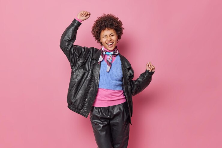 Foto grátis elegante mulher de cabelo encaracolado vestida com roupas de couro dança despreocupada tem expressão feliz esquece os problemas mantém os braços levantados se sente otimista isolado sobre fundo rosa pessoas e moda