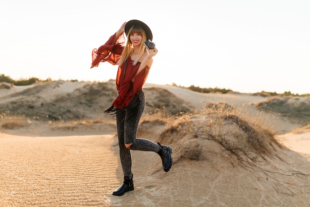 Elegante mulher posando no deserto, vestindo roupa de chapéu preto e boho