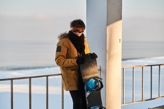 Em dia ensolarado, jovem snowbordist está posando para o fotógrafo com sua prancha de snowboard.