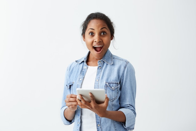 Emocional atônita jovem de pele escura na camisa azul, segurando nas mãos tablet, surpreso e chocado com notícias inesperadas positivas. Expressões e emoções do rosto humano