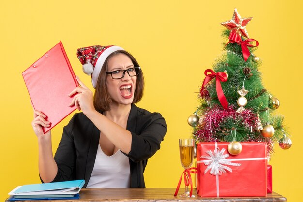 Emocional nervosa encantadora senhora de terno com chapéu de papai noel e óculos no escritório em amarelo isolado