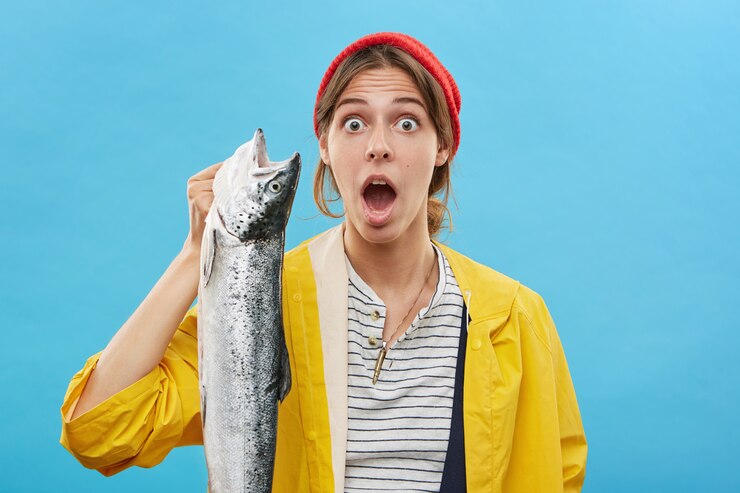Foto grátis emocional surpreendida jovem pescadora vestindo capa de chuva amarela e chapéu segurando um grande peixe na mão e olhando com a boca aberta, chocada com a boa captura. conceito de pesca