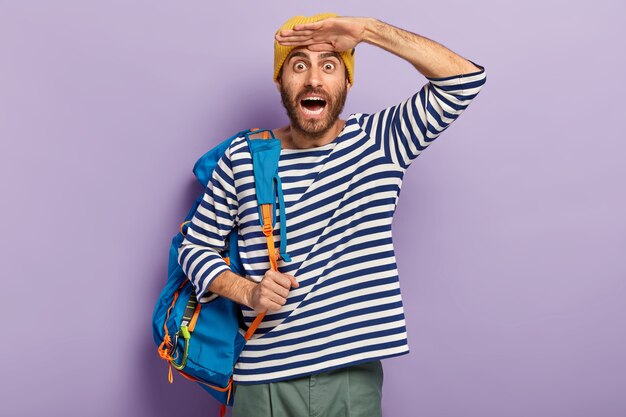 Emocional surpreso alegre jovem turista masculino mantém a palma da mão perto da testa, vestido com um suéter listrado, carregando uma mochila azul com coisas pessoais