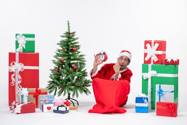 Emocionalmente louco surpreendeu o papai noel sentado no chão e mostrando o relógio perto de presentes e uma árvore de Natal decorada em fundo branco