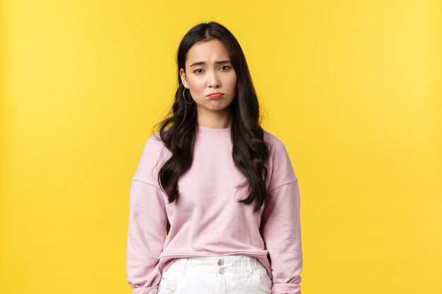 Emoções de pessoas, estilo de vida e conceito de moda. Menina coreana deprimida e triste, sombria fazendo beicinho, olhando para baixo em lixões, sentindo-se chateada e descontente, com um fundo amarelo de pé.