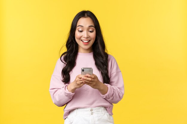 Emoções de pessoas, lazer do estilo de vida e conceito de beleza. Menina asiática surpresa e feliz recebe ótimas notícias pelo telefone, olhando para o display do celular com um sorriso surpreso, regozijando o fundo amarelo de pé.