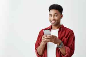 Foto grátis emoções positivas. close-up de jovem bonito homem de pele escura com penteado afro em camiseta branca e camisa vermelha, sorrindo com dentes, conversando com um amigo no smartphone