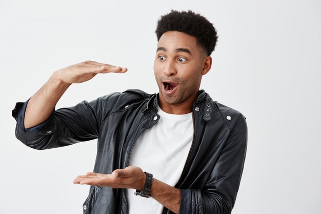 Emoções positivas. Copie o espaço. Retrato de jovem estudante masculino americano de pele bronzeada animado com cabelos cacheados em camiseta branca e jaqueta de couro gesticulando com as mãos, olhando de lado