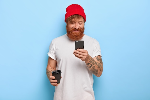 Emoções positivas e conceito de tecnologias modernas. Homem alegre e elegante usa chapéu vermelho e camiseta branca, conversa com amigos com barba ruiva pelo celular conectado à internet sem fio bebe café