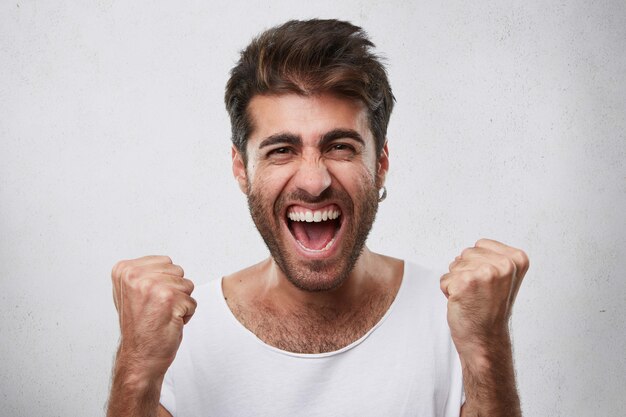 Emoções positivas, vitória, triunfo, conceito de felicidade. homem feliz com barba vestido com uma camiseta branca casual cerrando os punhos enquanto se alegra com sua vitória no jogo posar sobre o branco