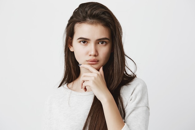 Emoções, sentimentos, reações e atitudes humanas. Mulher bonita em camiseta casual, com longos cabelos escuros e lisos, mantendo a mão no queixo em dúvida e suspeita, sentindo-se cético sobre algo