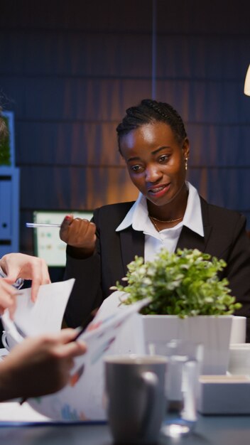 Empreendedor afro-americano focado em brainstorming de estratégia de empresa de negócios trabalhando ovetime na sala de reuniões à noite. Diversos colegas de trabalho multiétnicos analisando a papelada de apresentação de gerenciamento