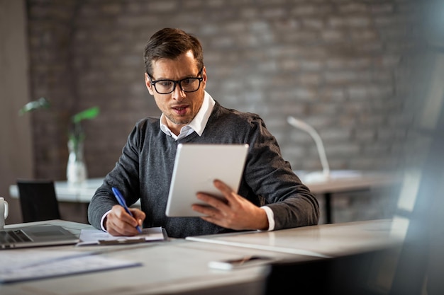 Empreendedor masculino escrevendo relatório de negócios enquanto usa tablet digital no escritório