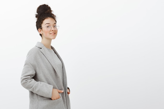 Empreendedora bem-sucedida, elegante, de casaco cinza e óculos, de pé meio virado com as mãos nos bolsos e sorrindo amigavelmente