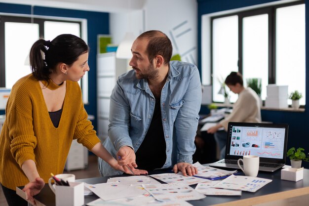 Empreendedores multiétnicos discutindo gráficos de dados no escritório olhando uns para os outros. Equipe diversificada de executivos que analisam relatórios financeiros da empresa a partir do computador. Arranque empresarial bem sucedido p