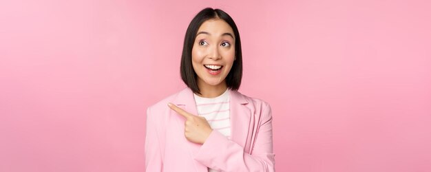 Empregada de escritório jovem empresária asiática entusiasmada, apontando o dedo para a esquerda, olhando para o anúncio de banner com um sorriso feliz, mostrando o fundo rosa do anúncio