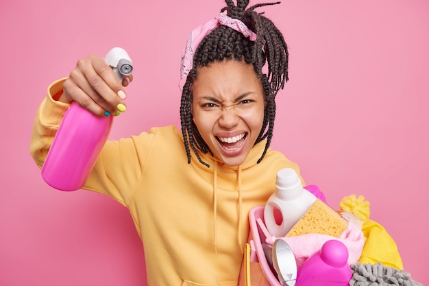 Foto grátis empregada doméstica irritada exclama, expressando emoções negativas com raiva segurando uma bacia cheia de roupa e um frasco de detergente