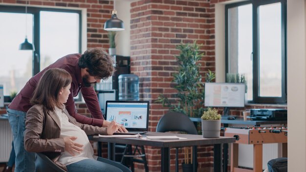 Empregada grávida trabalhando em um projeto de negócios com o laptop, falando com um homem sobre o desenvolvimento. mulher grávida e usando o computador para planejar a estratégia de marketing no escritório de inicialização.