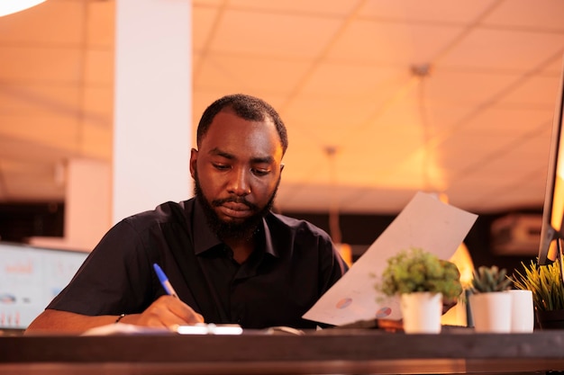 Empregado americano africano tomando notas sobre o contrato de inicialização, analisando a papelada com dados comerciais no escritório. trabalhar com documentos para pesquisar e encontrar soluções executivas.