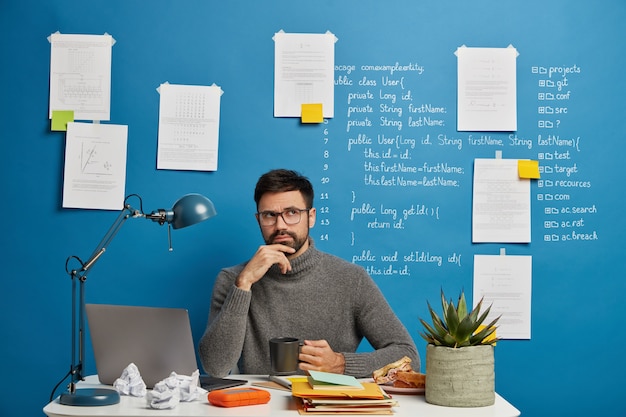 Empregado barbudo pensativo em gola olímpica casual, pensa em informações corporativas, segura a caneca de chá, posa no espaço de coworking, senta-se na frente do computador laptop contra o fundo azul.