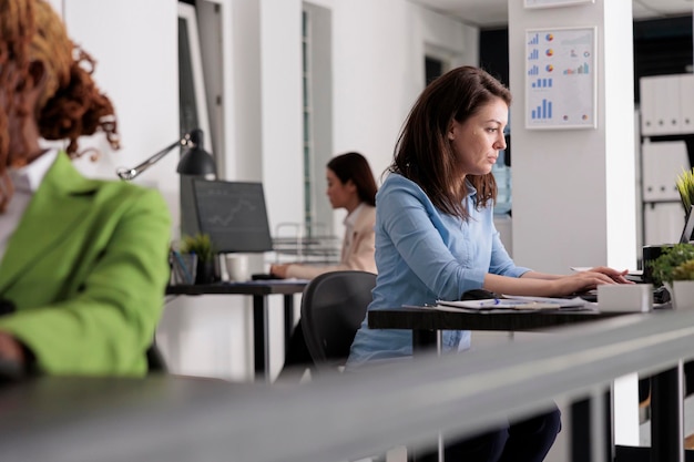 Empregado da empresa ocupada trabalhando na mesa no espaço de coworking, gerente corporativo no local de trabalho, vista lateral. Mulher séria atraente digitando no laptop no escritório moderno, foco seletivo