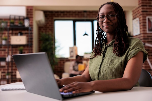 Foto grátis empregado da empresa trabalhando remotamente digitando casual sorrindo para a câmera enquanto o colega de quarto desfruta do tempo livre em casa. retrato de estudante afro-americano aprendendo usando computador portátil sentado na mesa.