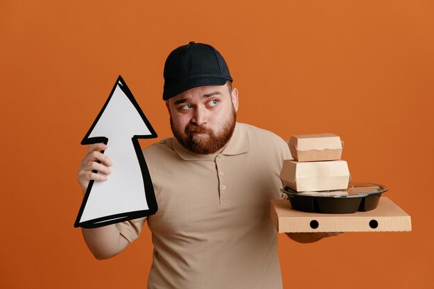 Empregado de entregador de boné preto e uniforme de camiseta em branco segurando a seta e recipientes de comida olhando de lado confuso com dúvidas sobre fundo laranja