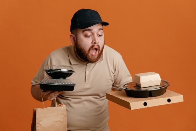 Empregado de entregador de boné preto e uniforme de camiseta em branco segurando recipientes de comida com saco de papel parecendo preocupado em pé sobre fundo laranja