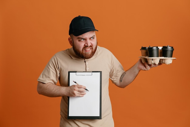 Empregado de entregador de boné preto e uniforme de camiseta em branco segurando xícaras de café e prancheta com caneta esperando uma assinatura olhando de lado descontente com cara feia em pé sobre fundo laranja