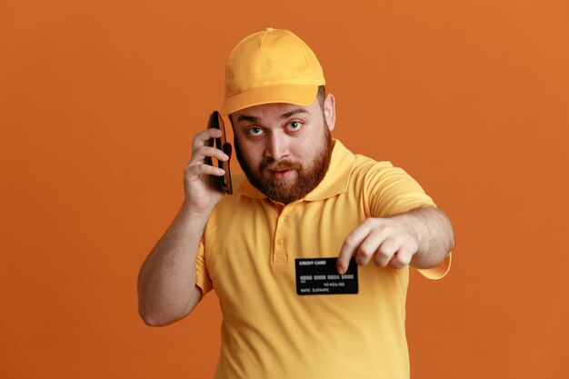 Empregado de entregador em uniforme de camiseta em branco de boné amarelo mostrando cartão de crédito falando no celular parecendo confiante em pé sobre fundo laranja