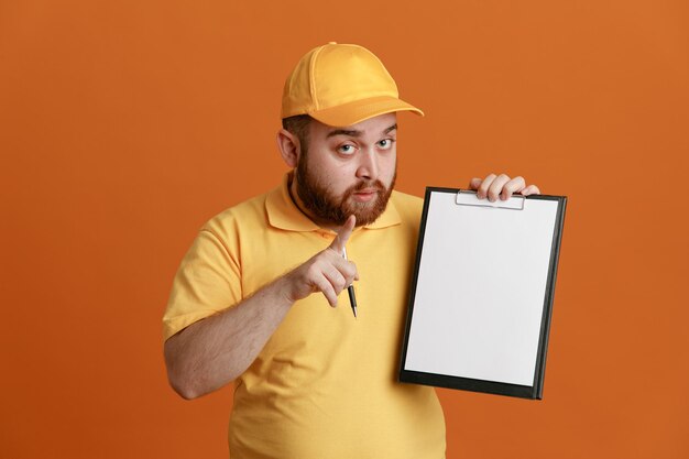 Empregado de entregador em uniforme de camiseta em branco de boné amarelo segurando a área de transferência e caneta apontando com o dedo indicador para a câmera sendo confiante em pé sobre fundo laranja