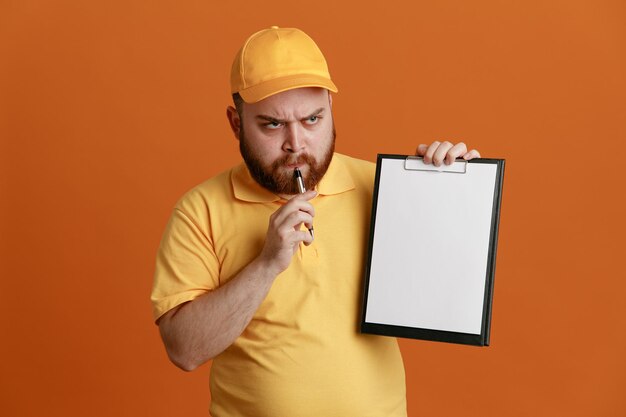 Empregado de entregador em uniforme de camiseta em branco de boné amarelo segurando a prancheta e caneta olhando confuso pensando em pé sobre fundo laranja