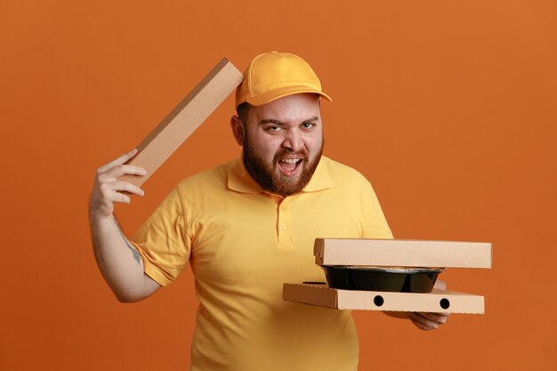 Empregado de entregador em uniforme de camiseta em branco de boné amarelo segurando recipientes de comida e caixa de pizza olhando para a câmera com expressão irritada em pé sobre fundo laranja