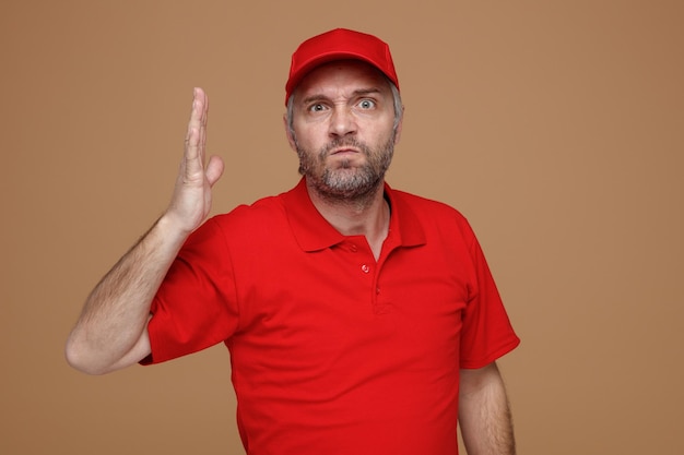 Empregado de entregador em uniforme de camiseta em branco de boné vermelho olhando para a câmera com raiva e frustrado, levantando o braço em desagrado em pé sobre fundo marrom