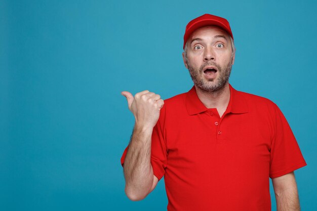 Empregado de entregador em uniforme de camiseta em branco de boné vermelho olhando para a câmera espantado e surpreso apontando com o polegar para o lado em pé sobre fundo azul