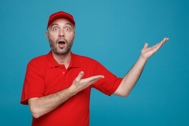 Empregado de entregador em uniforme de camiseta em branco de boné vermelho olhando para a câmera espantado e surpreso apresentando com os braços das mãos algo de pé sobre fundo azul