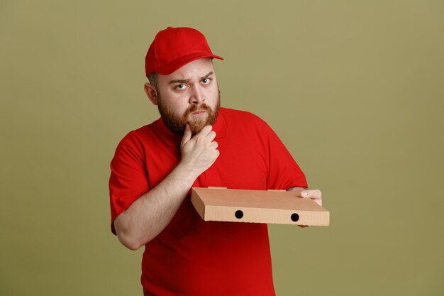 Empregado de entregador em uniforme de camiseta em branco de boné vermelho segurando a caixa de pizza olhando para a câmera com expressão pensativa com a mão no queixo pensando em pé sobre fundo verde