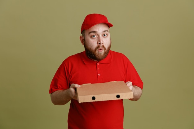 Empregado de entregador em uniforme de camiseta em branco de boné vermelho segurando a caixa de pizza olhando para a câmera feliz e surpreso em pé sobre fundo verde