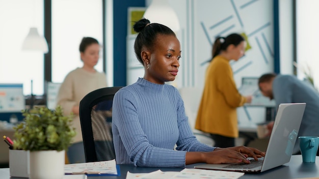 Empregado de inicialização americano africano sentado na mesa abrindo laptop e começando a digitar no teclado trabalhando em estatísticas de vendas. Mulher de negócios no escritório ocupado moderno começando a trabalhar pela manhã.