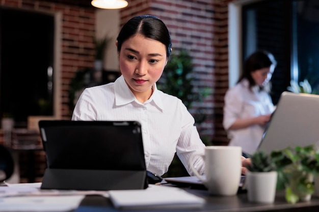 Empresária asiática com tablet moderno revisando a documentação contábil no espaço de trabalho do escritório. Gerente executivo trabalhando em dispositivo eletrônico touchscreen enquanto desenvolve a estratégia de marketing.